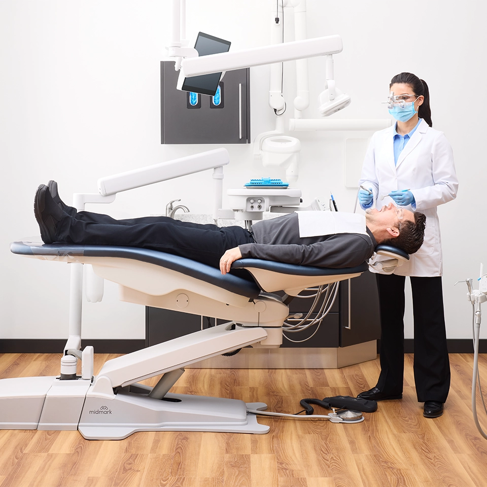 A Midmark Dental operatory featuring a Midmark Operatory chair, where a masked dentist wearing protective glasses stands beside a patient reclining in a modern dental chair under overhead exam lights in a clean, white treatment room.