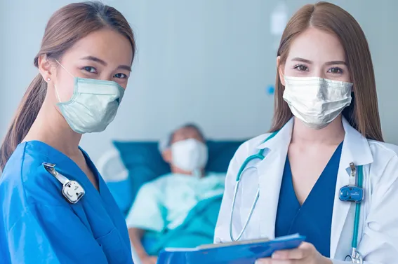 A nurse in blue scrubs and a doctor in a white lab coat wear surgical masks and RTLS badges while reviewing a patient chart in front of a patient bed.