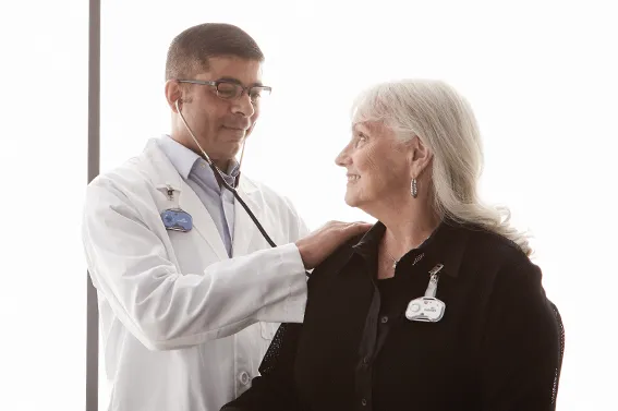 A doctor wearing a blue Midmark RTLS badge uses a stethoscope to examine an elderly female patient who is also wearing a white wearable clip-on RTLS asset tag.
