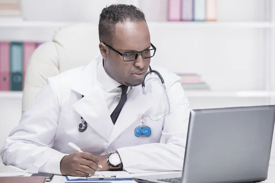 A male doctor wearing a white lab coat, tie, and a blue RTLS badge looks intently at a laptop screen while writing notes at a desk workspace.
