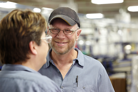 A smiling Midmark service technician wearing a baseball cap and glasses talks with a colleague in a brightly lit manufacturing facility.