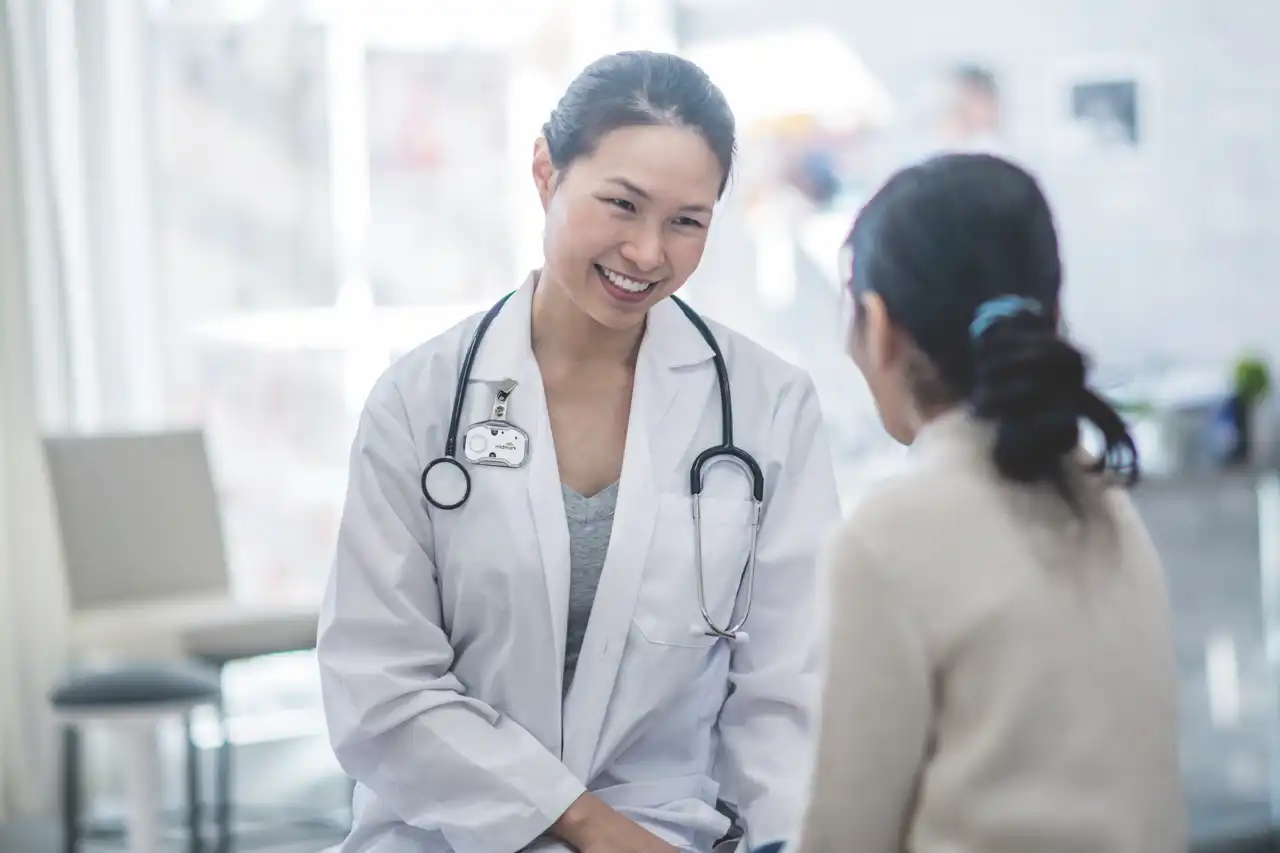 Woman with dark hair, white coat and stethoscope smiling at a woman with dark hair, pulled back