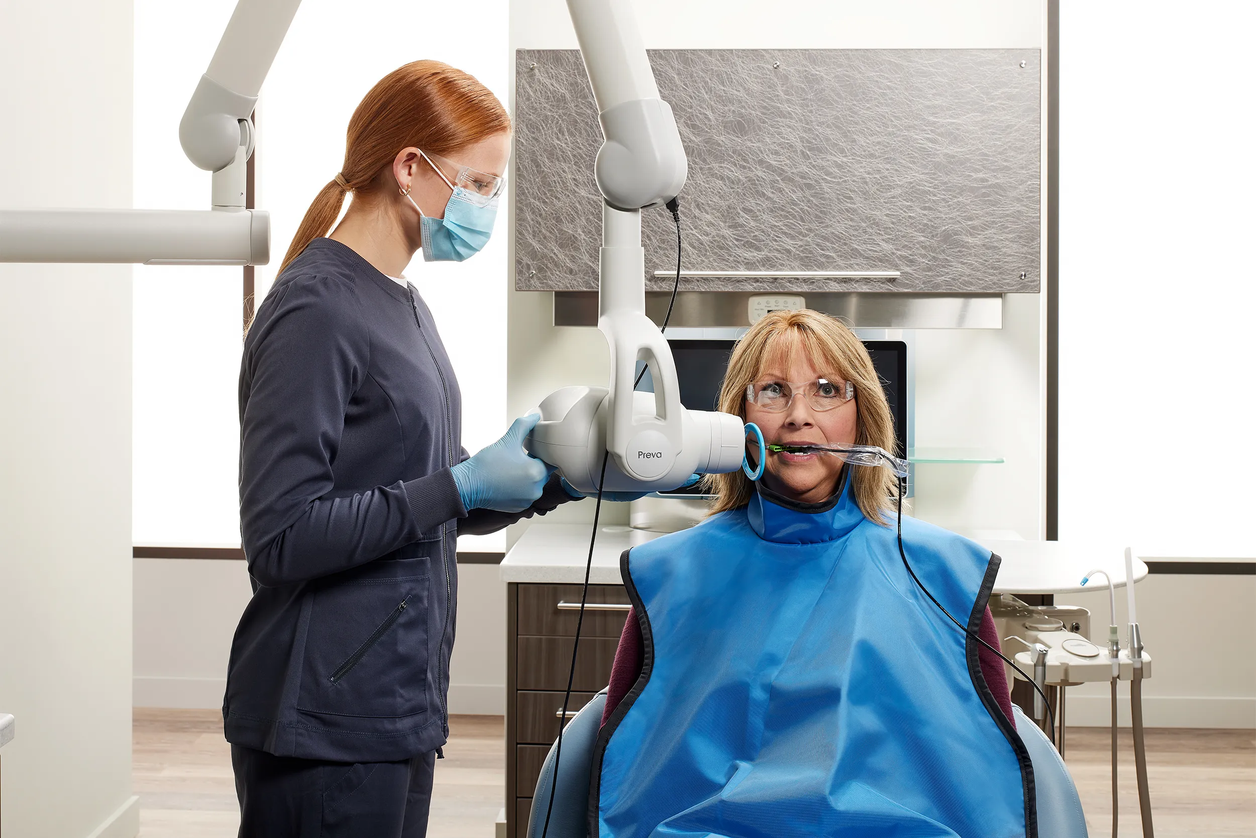 Dental professional positioning a dental X-ray unit for intraoral imaging while patient wears protective lead apron..