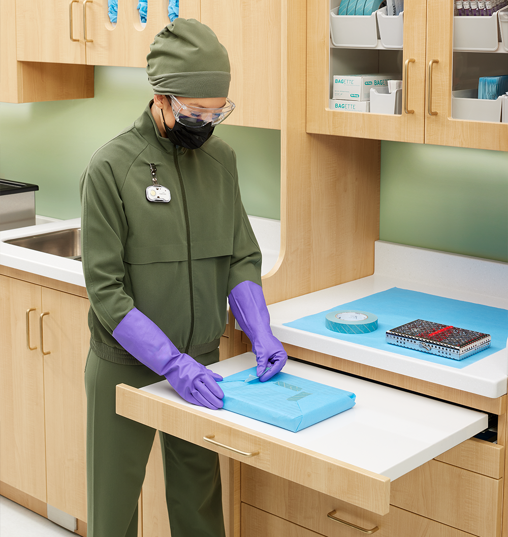 Healthcare worker wearing gloves wraps a sterilization cassette on a clean instrument processing work surface.
